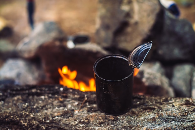 Heating tea in metal mug on bonfire with large firewood. Tea drinking in open air. Active outdoor recreation. Camping in dusk. Romantic warm atmosphere in twilight on nature. Active rest.