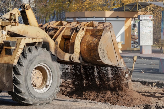 A heavy front loader transports earth in a bucket and grades the road. Road repair in the center of a modern city in autumn. Heavy construction machinery for moving soil.