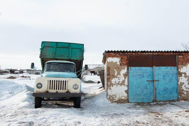 Un vecchio pesante autocarro con cassone ribaltabile blu è parcheggiato accanto a un edificio in mezzo alla neve bianca, in attesa che inizi il carico. consegna della merce in inverno