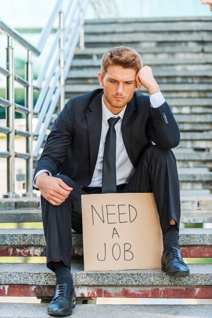 Foto heb een baan nodig. depressieve jongeman in formalwear met poster met sms-bericht terwijl hij op de trap zit