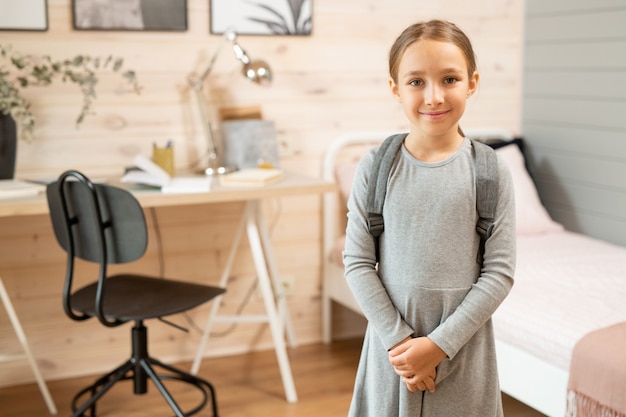 Foto hedendaags gelukkig schoolmeisje in grijze jurk die tegen haar bed staat, tafel bij houten muur en zwarte plastic stoel