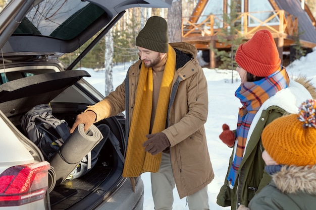 Hedendaags jong gezin van drie in warme winterkleding die hun bagage uit de kofferbak halen terwijl ze achter de auto staan tijdens het reizen