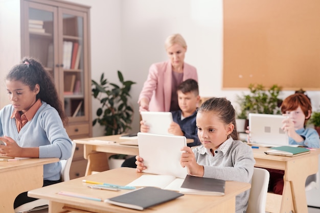 Hedendaags schoolmeisje en haar klasgenoten met touchpads die bij bureaus zitten en individueel werken terwijl de leraar bij een van hen staat