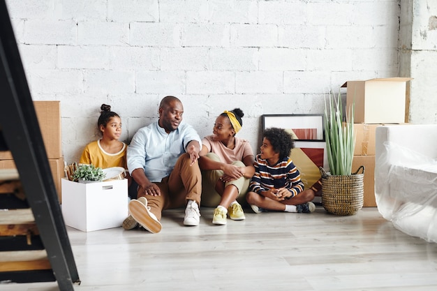 Hedendaagse afrikaanse familie van vader, moeder, zoontje en tienerdochter zittend op de vloer tegen een wit geschilderde muur en praten