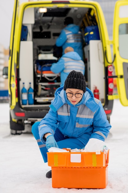 Hedendaagse jonge paramedicus in blauwe werkkleding en handschoenen op zoek naar medicijnen in EHBO-kit buitenshuis