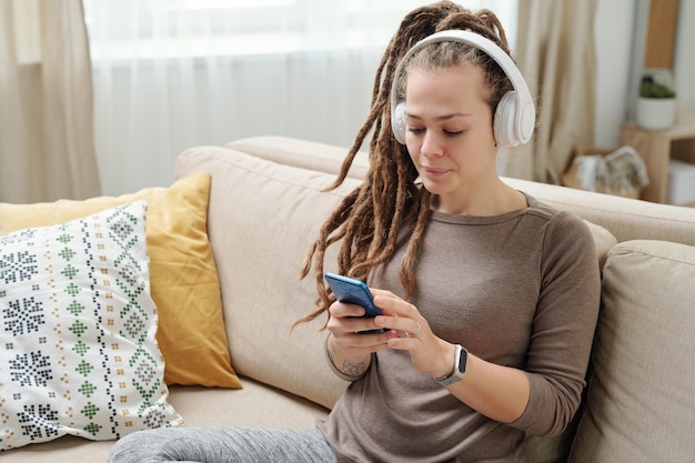 Hedendaagse jonge vrouw met dreadlocks luisteren naar muziek in hoofdtelefoons en scrollen in smartphone op Bank