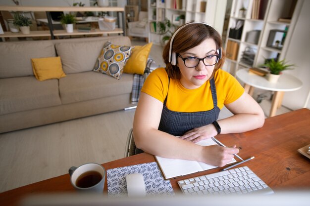 Hedendaagse jonge vrouwelijke student in koptelefoon uitschakelen computerscherm kijken en notities maken tijdens het volgen van een online cursus