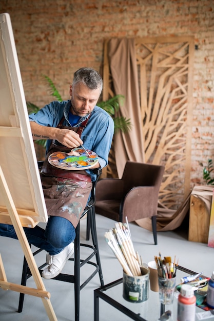 Hedendaagse schilder in vrijetijdskleding en schort die kleuren op palet mengt terwijl hij voor ezel in studio zit