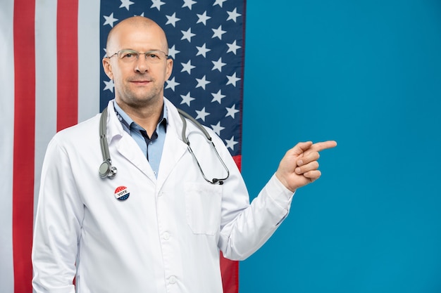 Hedendaagse volwassen Amerikaanse arts met een stethoscoop op de nek en insignes op de borst die naar voren wijzen terwijl hij tegen de vlag staat