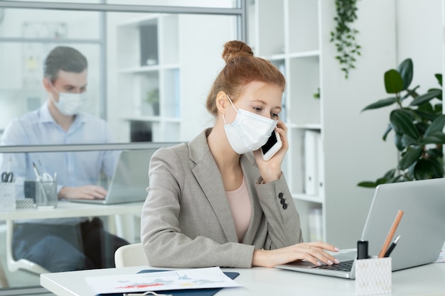Hedendaagse zakenvrouw met beschermend masker die naar een laptopscherm kijkt terwijl ze met gegevens werkt en op kantoor telefoneert tegen haar collega