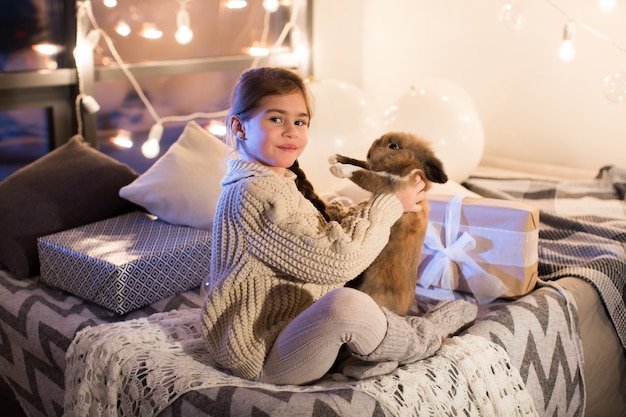 Heel mooi en charmant klein donkerharig meisje in witte trui heeft een levend konijn in het interieur van het huis. Nieuwjaar. Kerstmis. Haas. Studio.