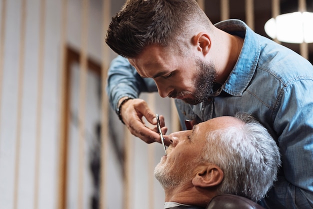 Heel voorzichtig. Zijaanzicht van de jonge knappe bebaarde kapper permanent en snor knippen