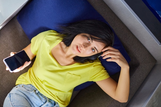 Photo heerful female in casual clothes with headphones and smartphone, listening to podcast on sofa at home.