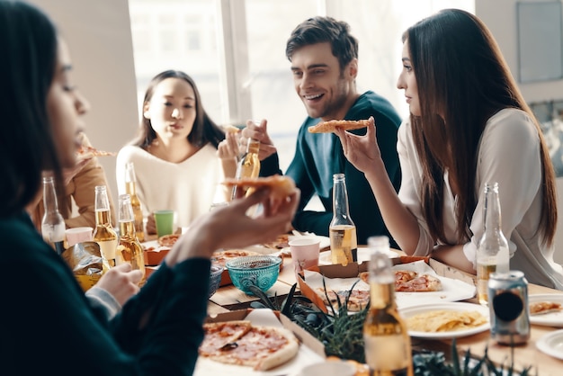 Heerlijk eten en geweldige mensen. Groep jongeren in vrijetijdskleding die pizza eet en glimlacht terwijl ze binnen een etentje hebben