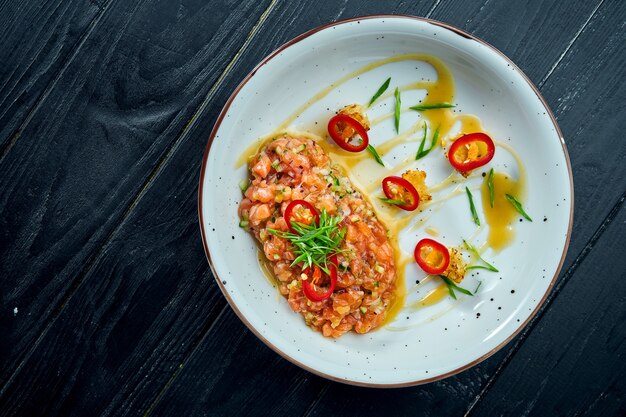 Heerlijk voorgerecht - zalmtartaar met avocado en hete chilipeper op een witte plaat op zwarte houten ondergrond.