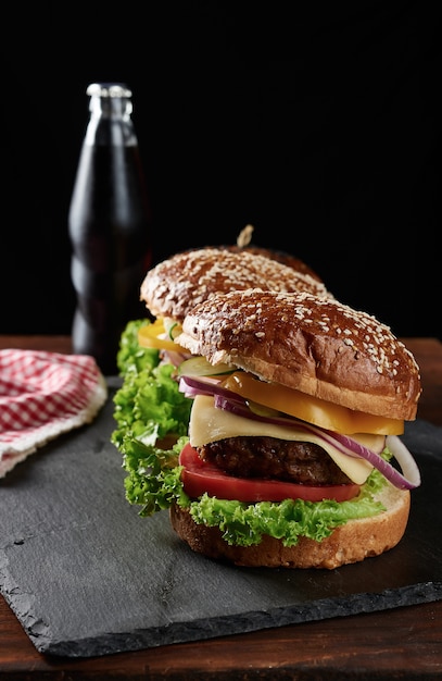 Heerlijke burgers met gebakken runderkotelet en uien, tomaat en krokant wit tarwebloembroodje met sesamzaadjes. Fastfood op een houten bord, achter een flesje met een drankje