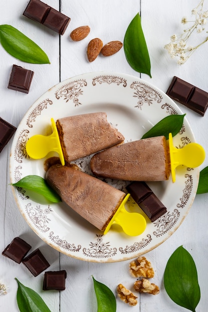 Heerlijke en verfrissende chocolade-ijslolly's in een vintage bord op een witte houten tafel bedekt met stukjes chocolade, noten, amandelen en bladeren uit de natuur