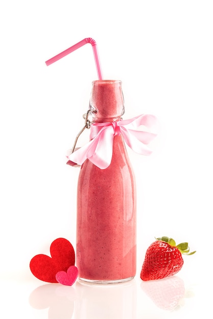 Heerlijke en voedzame smoothie gemaakt van verse aardbeien in een glazen fles met een rietje, een roze strik en stoffen hartjes. Cadeau om de geliefden te geven en te delen op Valentijnsdag. Witte achtergrond.