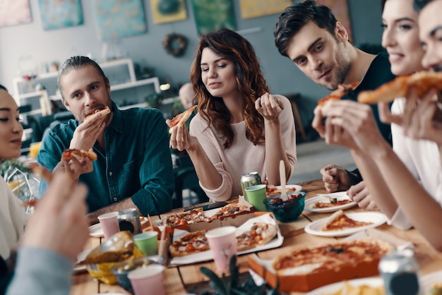 Heerlijke maaltijd delen. Groep jongeren in vrijetijdskleding die pizza eet en glimlacht terwijl ze binnen een etentje hebben