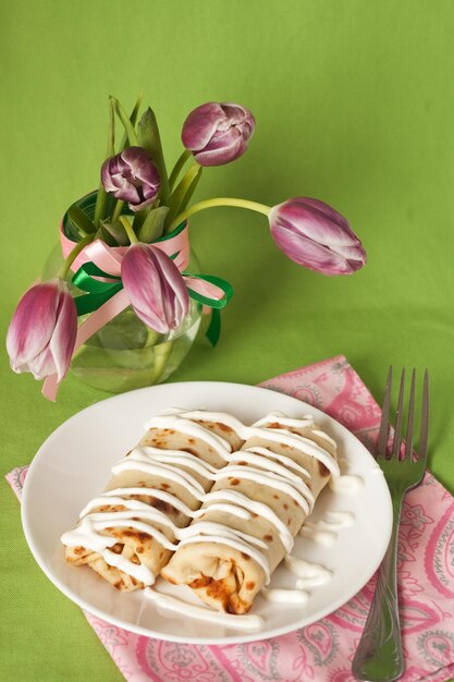 Heerlijke pannenkoeken met zure room op een witte plaat naast een boeket tulpen Groene achtergrond