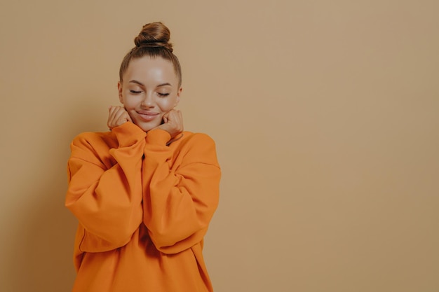 Heerlijke vrouw in oranje sweatshirt glimlachend met gesloten ogen, handen in de buurt van het gezicht houdend en dromend