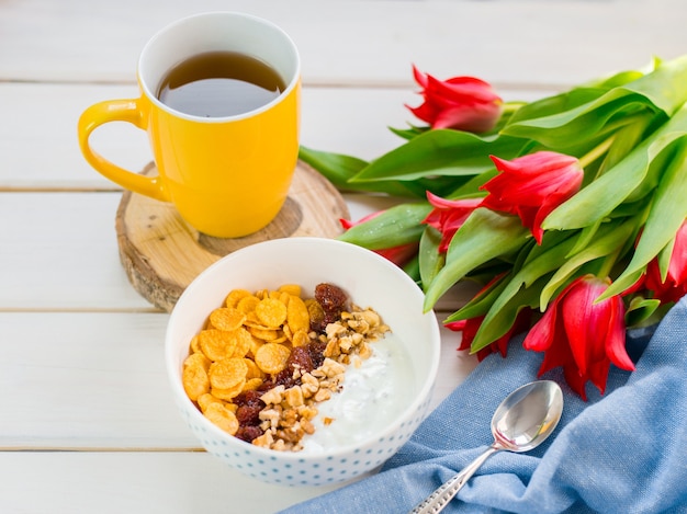 Heerlijke yoghurtkom met cornflakes, noten en jam op een witte houten lijst. Gezond en biologisch voedingsconcept. Tulpen met kopje thee en ontbijt.