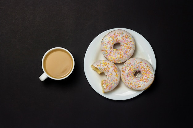 Heerlijke, zoete, verse donuts op een witte plaat, een kopje koffie, zwarte achtergrond. concept van ontbijt, fast food, coffeeshop, bakkerij. plat lag, bovenaanzicht.