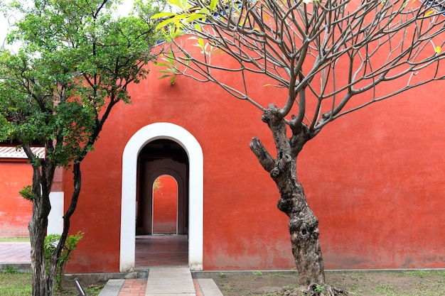 Heldere rode muur van Chinese tempel met gebogen deur, de oude bomen ernaast.