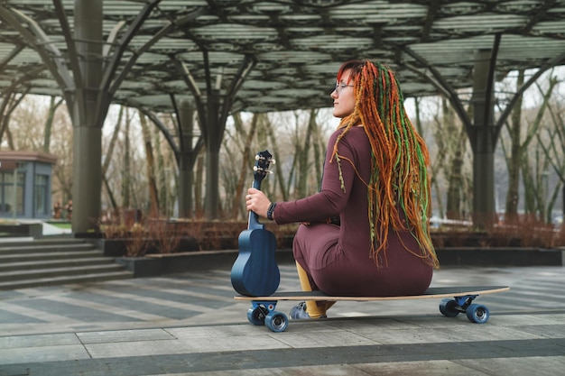 Heldere vrouw zit op een longboard op de weg en speelt gitaar ukulele op de weg