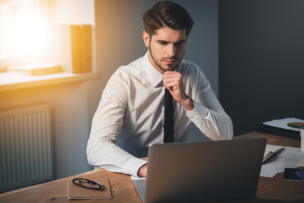 Helemaal geconcentreerd. Peinzende jonge knappe man die zijn laptop gebruikt en zijn hand op zijn kin houdt