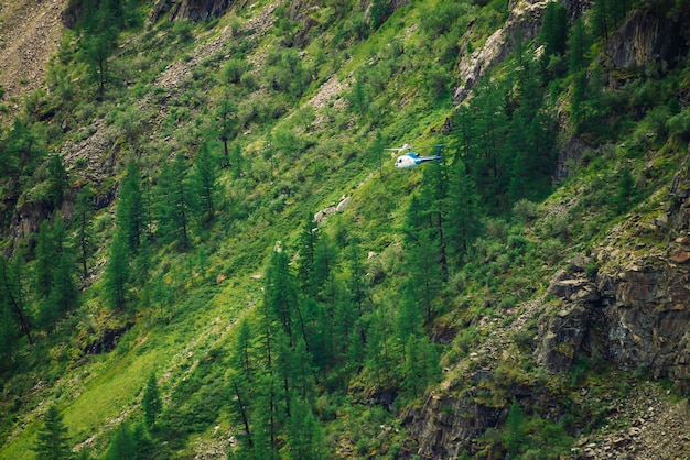 Helicopter in air against background of giant green rock covered with vegetation in tropical. Rescue mission in mountains. Aerial tourism in warm climate. Trees and grass on mountainside.