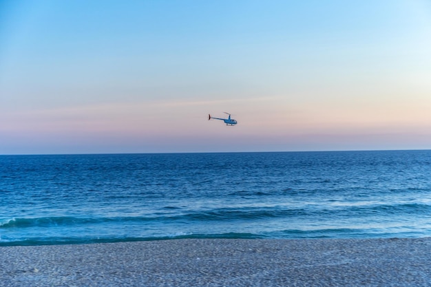 A helicopter flies over the ocean near a sandy beach