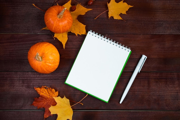 こんにちは秋のコンセプト落ちた黄色の葉カボチャと木製の背景に空白のメモ帳