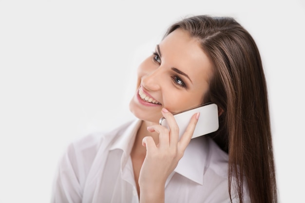 Фото Привет! красивая молодая женщина в белой рубашке разговаривает по мобильному телефону и улыбается, пока изолирована на белом