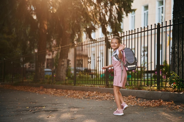 Foto ciao gesto della mano la scolaressa con lo zaino è all'aperto