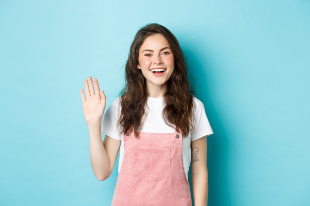 Hello nice to meet you. Beautiful friendly girl waving hand to say hi, looking happy and smiling while greeting you, giving warm welcome, standing over blue background