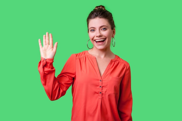 Hello. Portrait of charming woman with bun hairstyle, big earrings and in red blouse standing, waving hand and saying Hi to camera with toothy smile. indoor studio shot isolated on green background