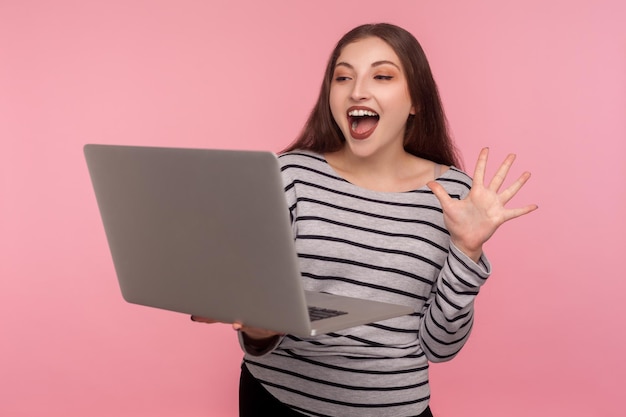 Hello. Portrait of friendly woman in striped sweatshirt waving hand at laptop screen, saying hello, communicating via video call, online conference. indoor studio shot isolated on pink background