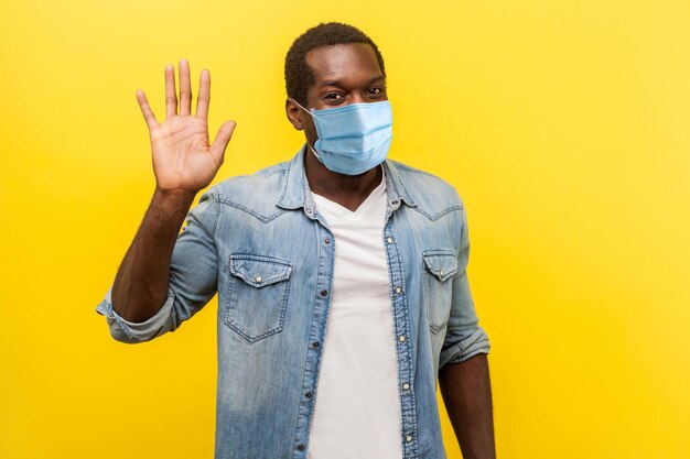 Hello Portrait of positive handsome man with medical mask with rolled up sleeves smiling friendly and waving hand saying hi welcoming gesture indoor studio shot isolated on yellow background