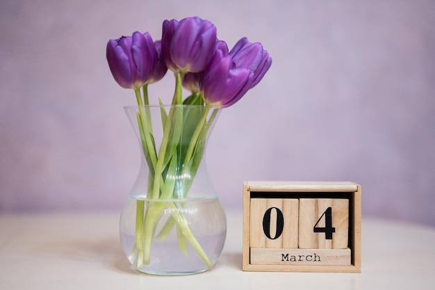 Hello spring Wooden cubic calendar with date of 4 March surrounded with bouquet of purple tulips with green leaves in glass vase