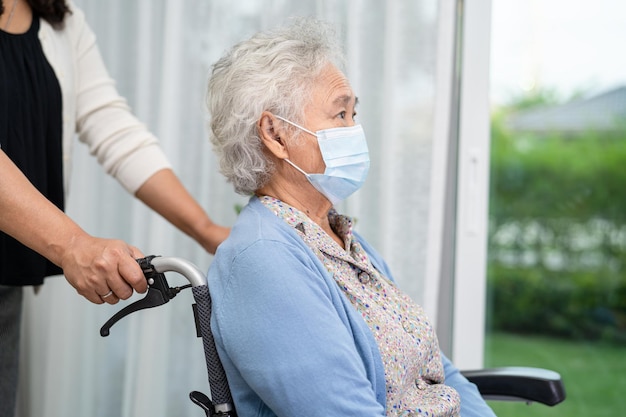 安全感染Covid-19コロナウイルスを保護するために車椅子に座ってフェイスマスクを着用しているアジアの年配の老婦人女性を助けてください。