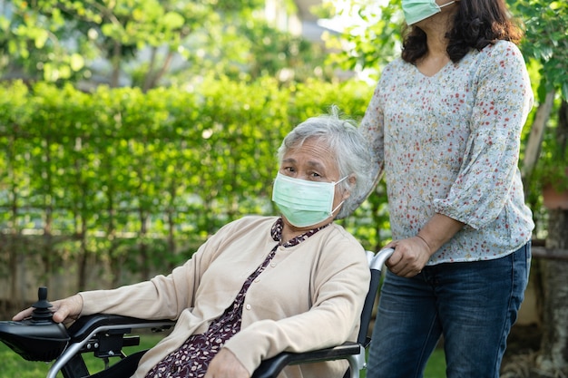 公園でコロナウイルスを保護するために電動車椅子とマスクを身に着けているアジアの年配の女性を助けます
