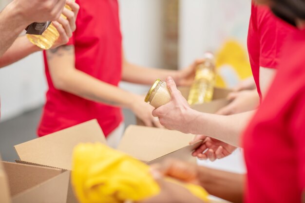 Help, liefdadigheid. Handen van jonge mensen in bijpassende rode t-shirts die voedsel en kleding verpakken in kartonnen dozen in een liefdadigheidsorganisatie