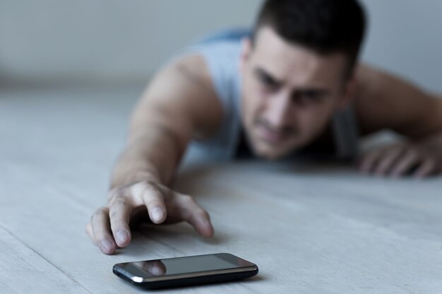 Help me! Frustrated young man lying on the floor and stretching hand to the mobile phone