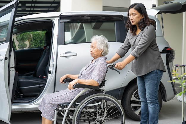 車椅子に座っているアジアのシニアまたは高齢の老婦人女性患者が彼女の車に健康で強力な医療コンセプトを取得する準備をするのを助け、サポートする