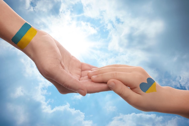 Photo help for ukraine woman and child holding hands with drawings of ukrainian flag and beautiful blue sky on background closeup