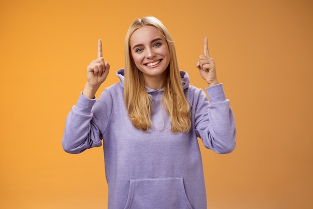 Helpful friendly-looking supportive charming female friend pointing up showing awesome new product advertisement suggesting check out tilting head gladly smiling help out, orange background.