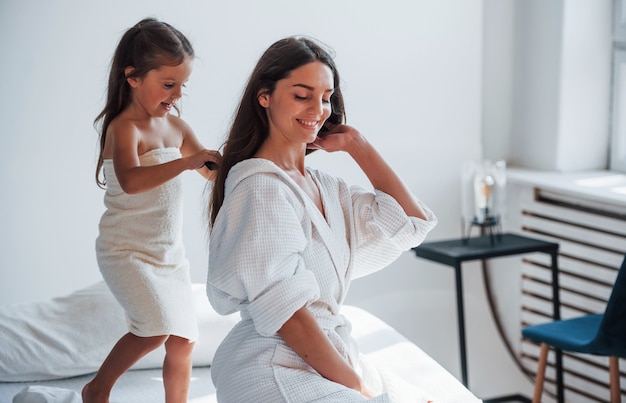 Helping to made hairstyle. Young mother with her daugher have beauty day indoors in white room.