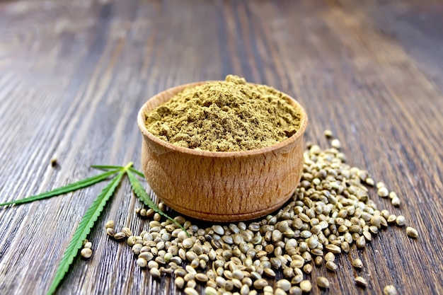 Hemp flour in a bowl, corn and green leaves of hemp on a background of wooden planks