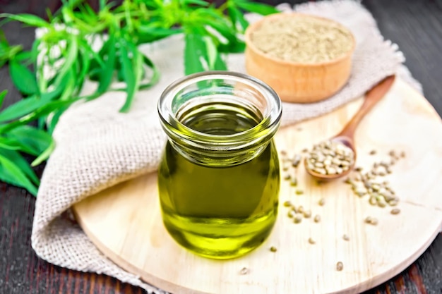 Hemp oil in a glass jar, grain in a spoon and flour in a bowl, leaves and stalks of cannabis, a napkin of burlap on the background of a dark wooden board